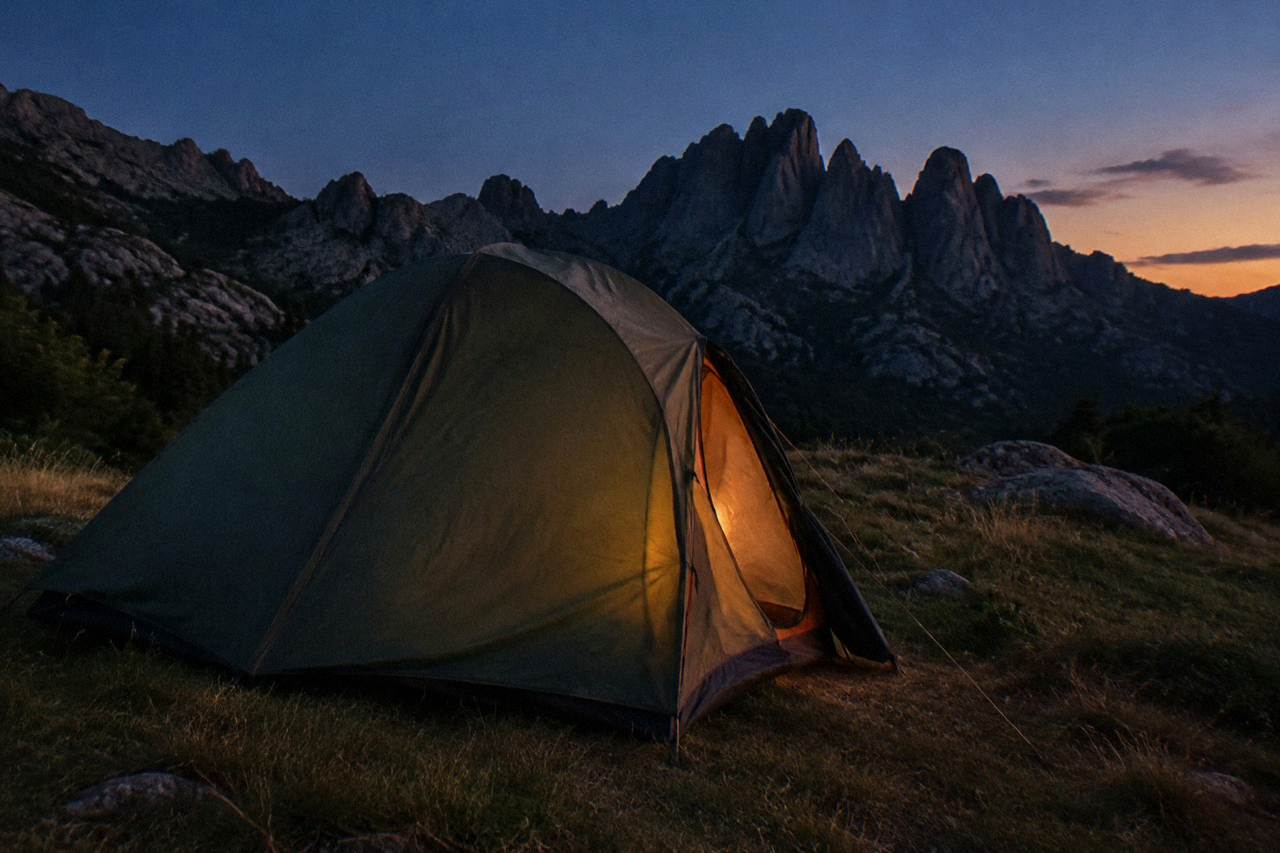 A tent pitched on a grassy mountain plateau with granite peaks behind it at dusk