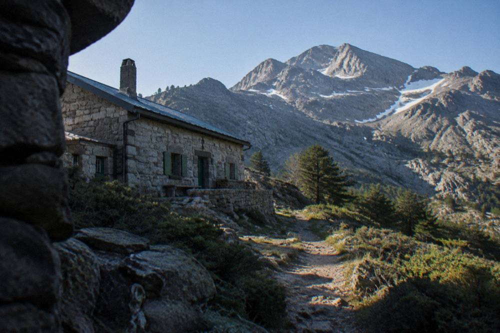A stone refuge building beside a mountain path with morning light on the ridgeline