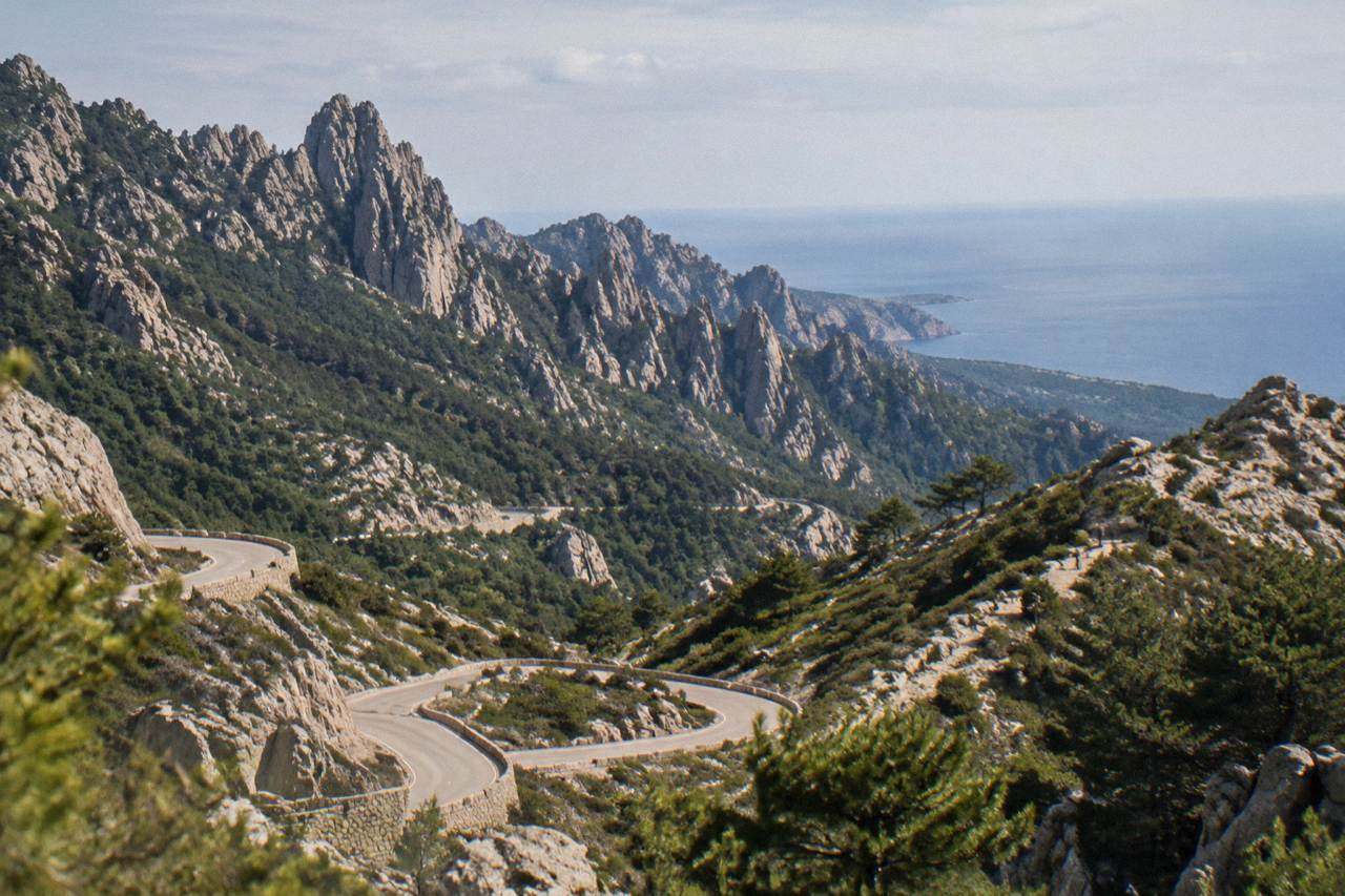 A winding mountain road with hairpin bends climbing through rocky Corsican terrain toward the sea