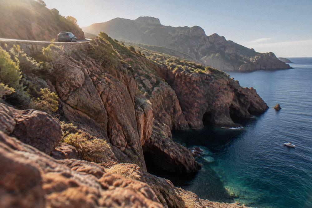 A car on a narrow coast road above red granite cliffs with the sea beyond