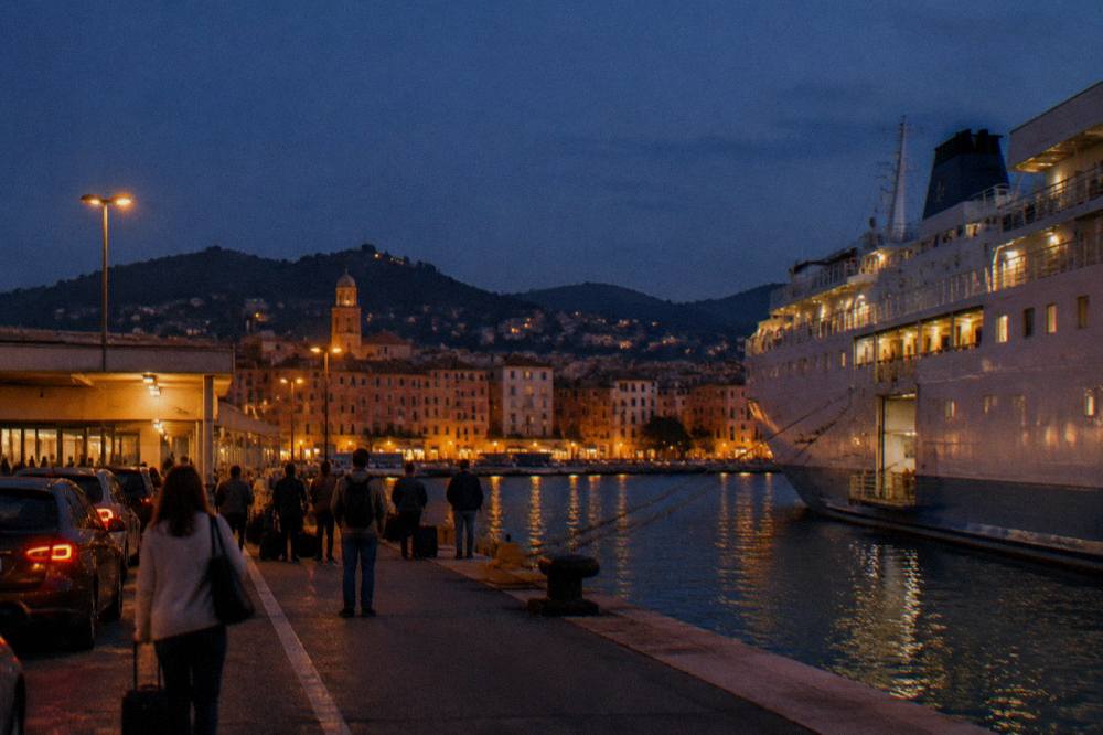 A ferry terminal quay at dusk with a moored ship and passengers waiting beside cars