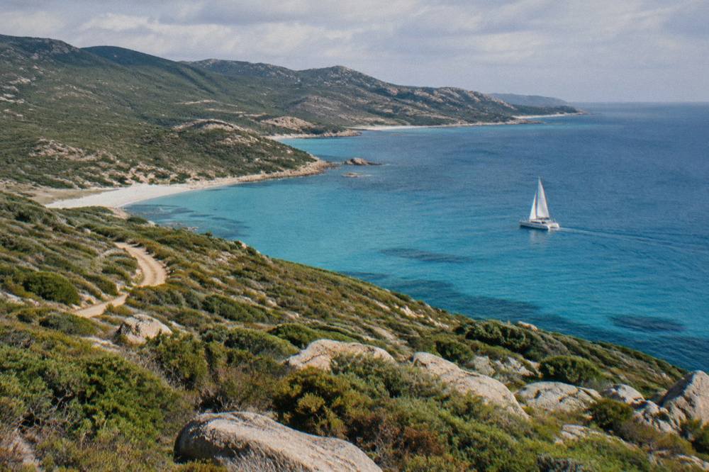 A white catamaran sailing along the Agriates coastline of northern Corsica with empty beaches and maquis-covered hills