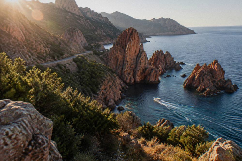 A small boat cruising along red granite rock formations above clear water on the Corsican west coast