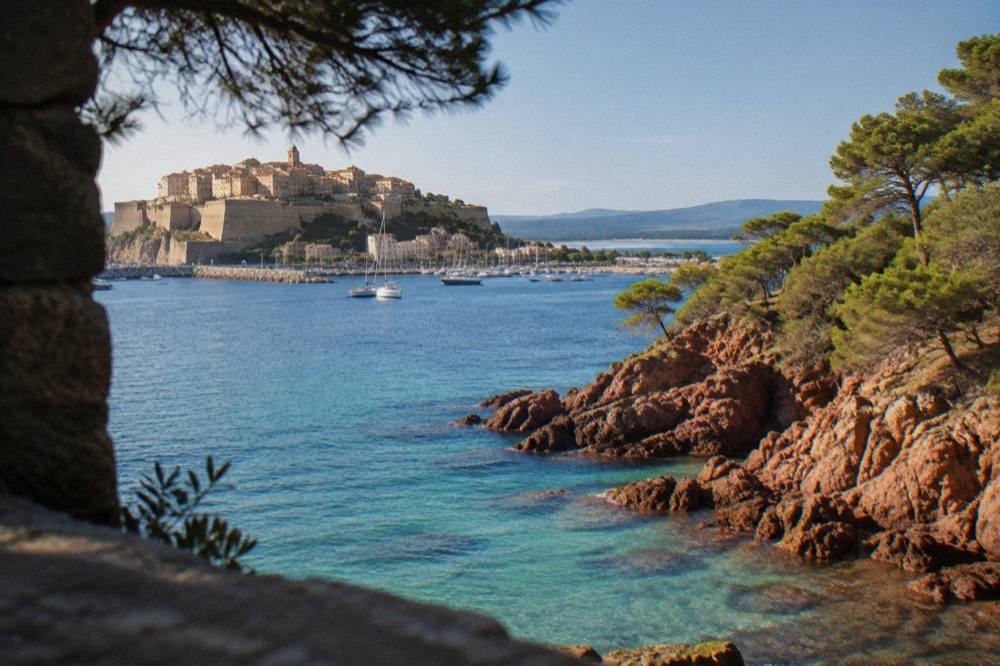 A rocky point with red granite formations and pine trees above turquoise water on the coast near Porto-Vecchio