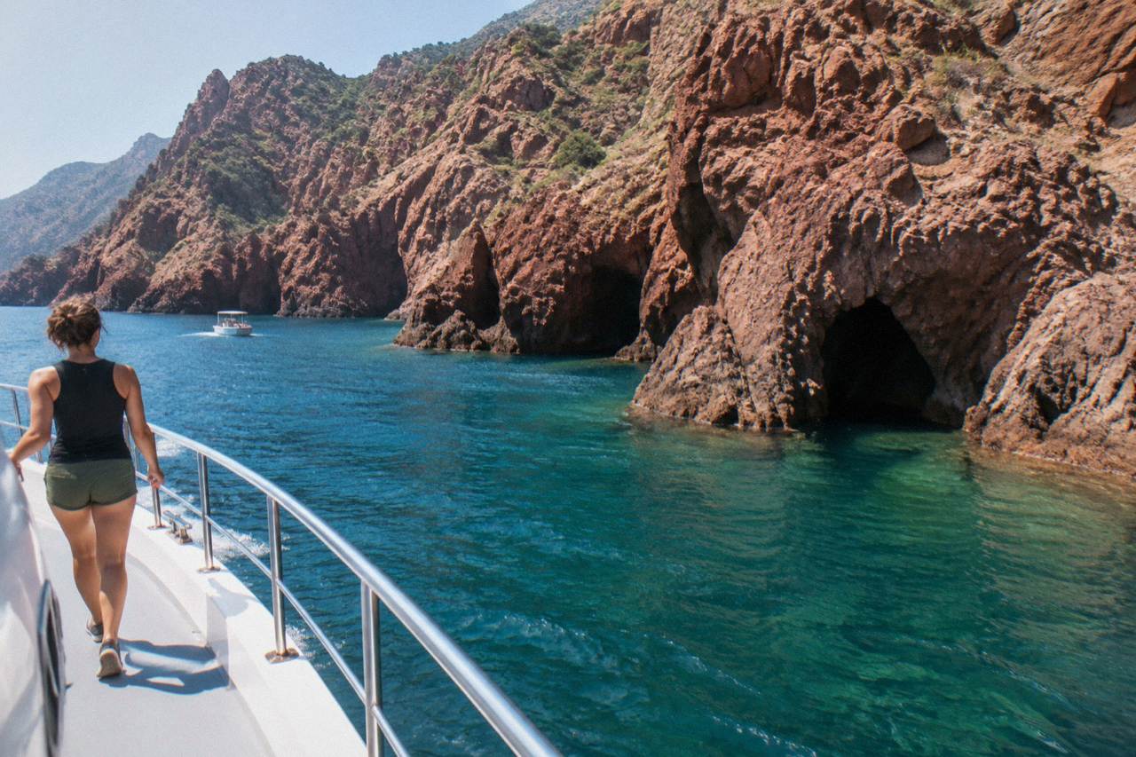 A boat cruising along red rocky cliffs above clear water at the Scandola reserve in Corsica