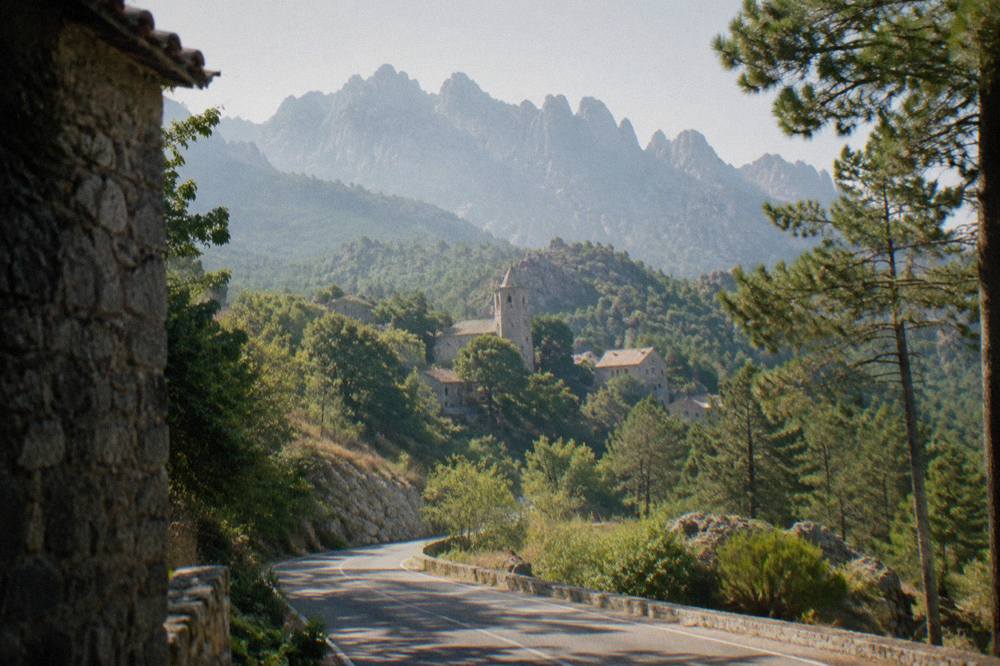 A mountain road winding through pine trees toward granite summits in the Corsican interior
