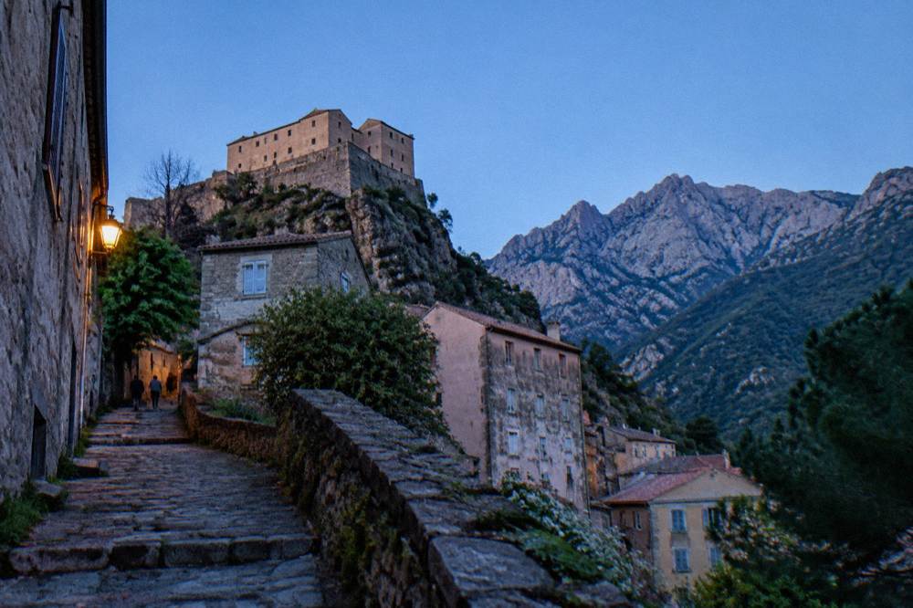 Stone houses below a citadel on a rock with high mountains behind