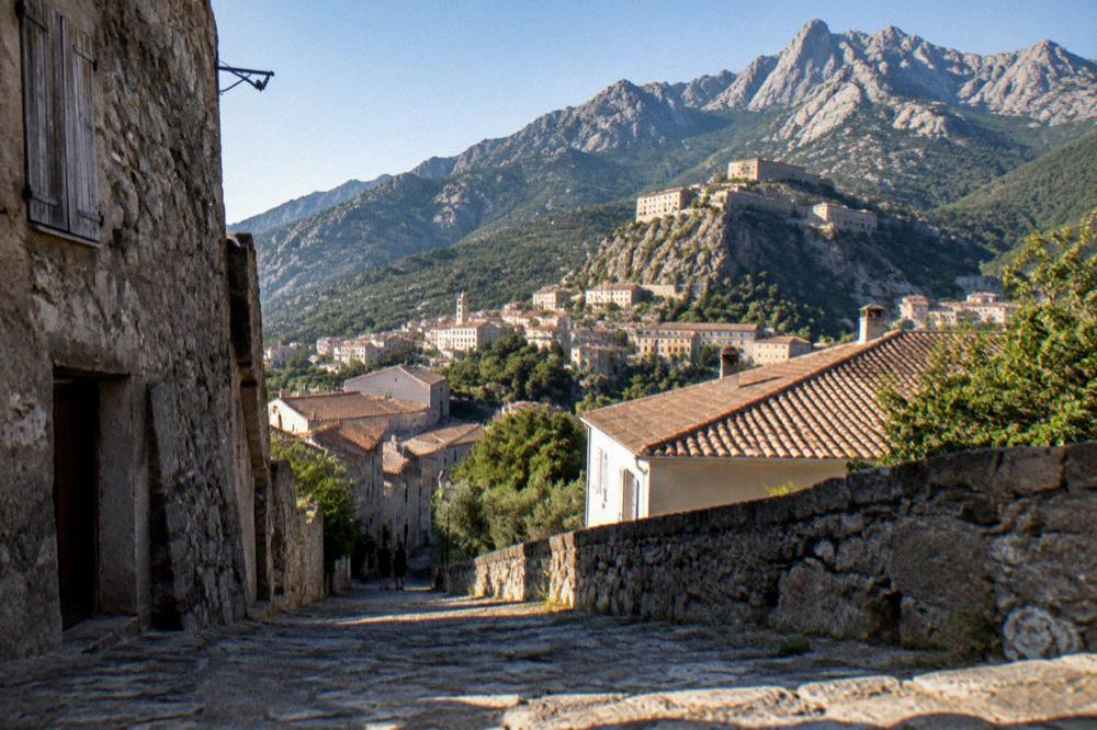 A mountain town with terracotta roofs under a clear sky with high peaks behind