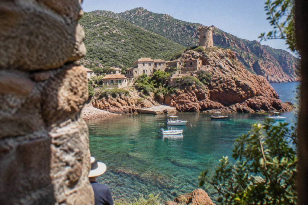 A red rocky Corsican coastline with clear water and green maquis hills behind