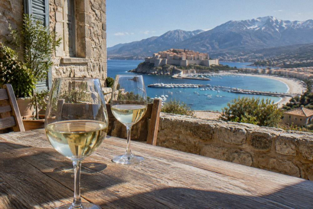 White wine glasses on a wooden table outside a stone building in the Corsican hills