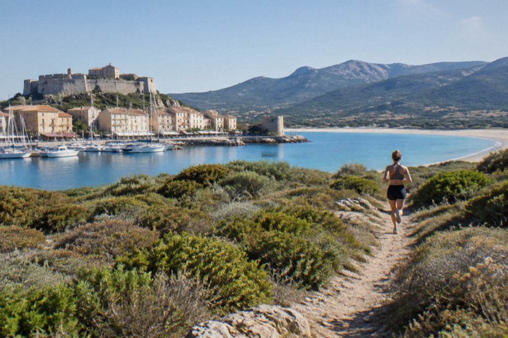 A footpath crossing low Mediterranean scrub toward a distant sandy bay