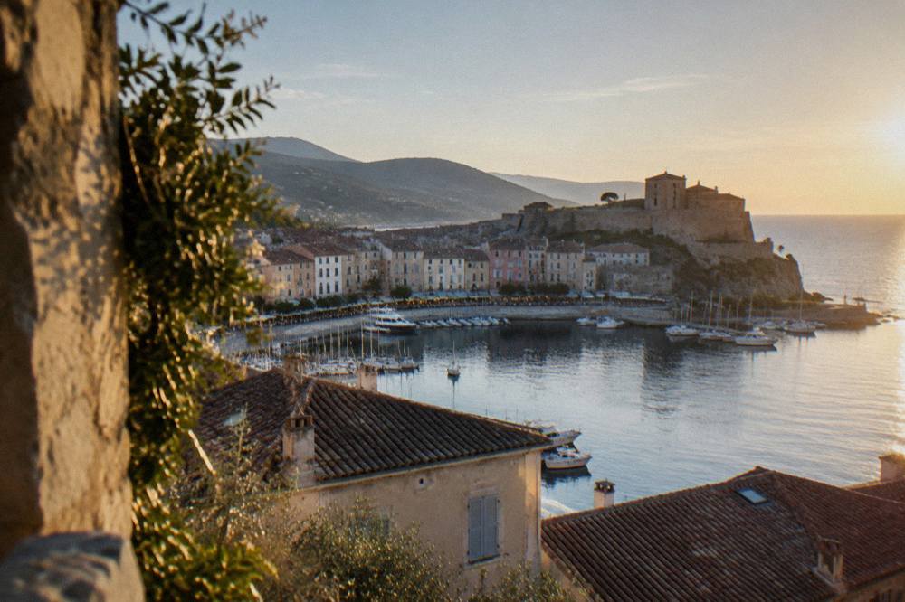 An evening view over tiled rooftops of a small town with a stone citadel tower and the sea beyond