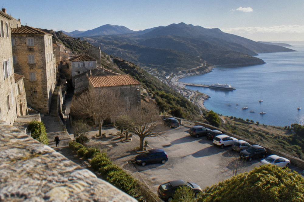 A car park above a gulf with a ferry and small boats at a jetty below
