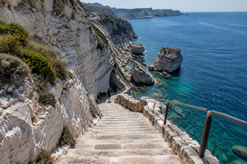A narrow stone stairway descending the cliff face towards clear water far below