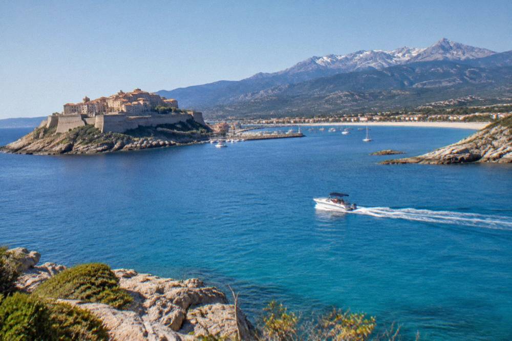A boat cutting across clear water in the Gulf of Calvi with rocky Corsican headlands behind