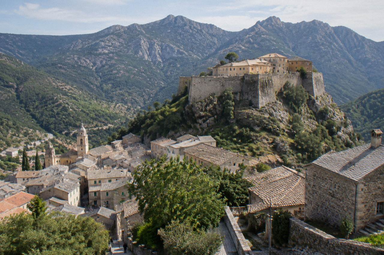 A grey stone citadel on a rocky outcrop above a mountain town under heavy clouds