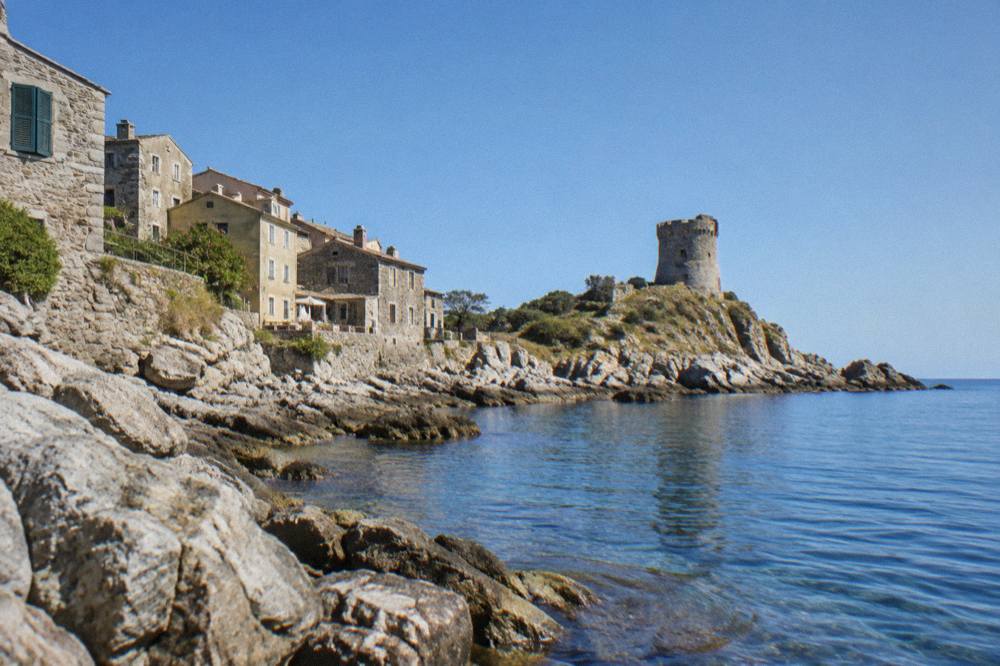 A rocky peninsula reaching into calm blue Mediterranean water with a stone tower on the headland