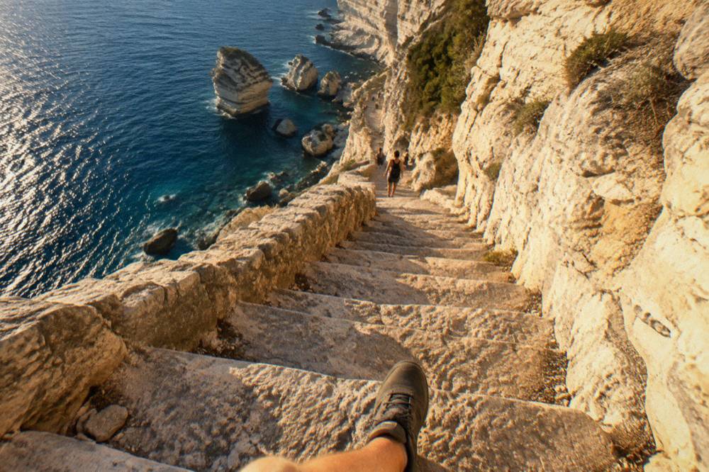 A person in walking shoes on worn stone steps with the sea visible far below