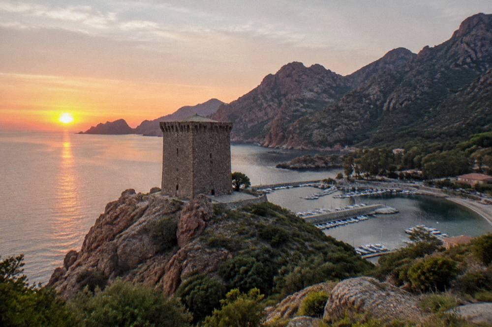 The sun setting over dark red rock formations along a calm sea