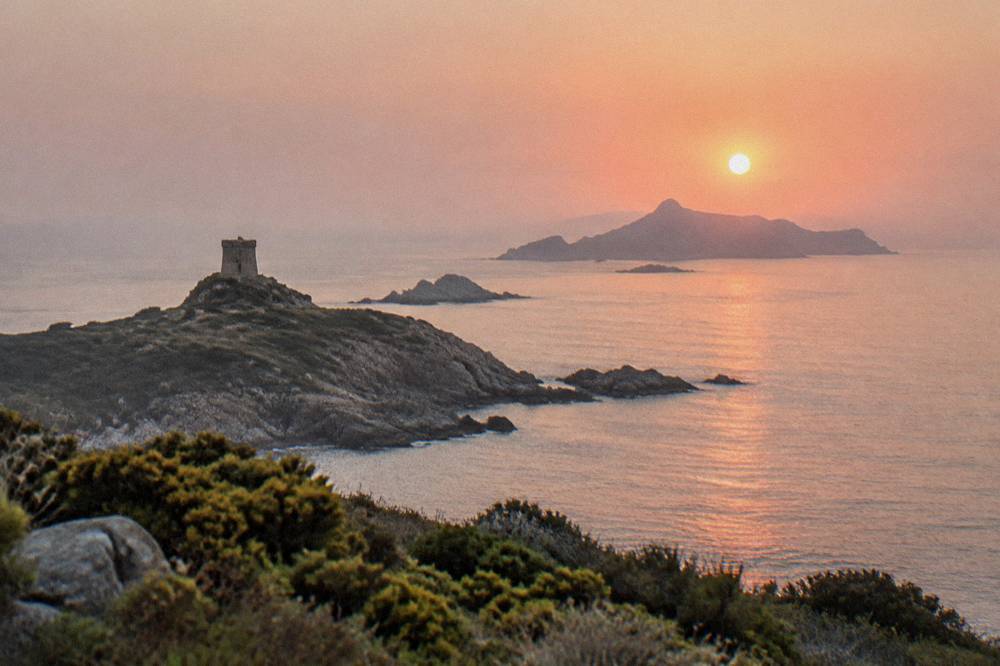 The sun low over the sea horizon behind a rocky island silhouette seen from the water