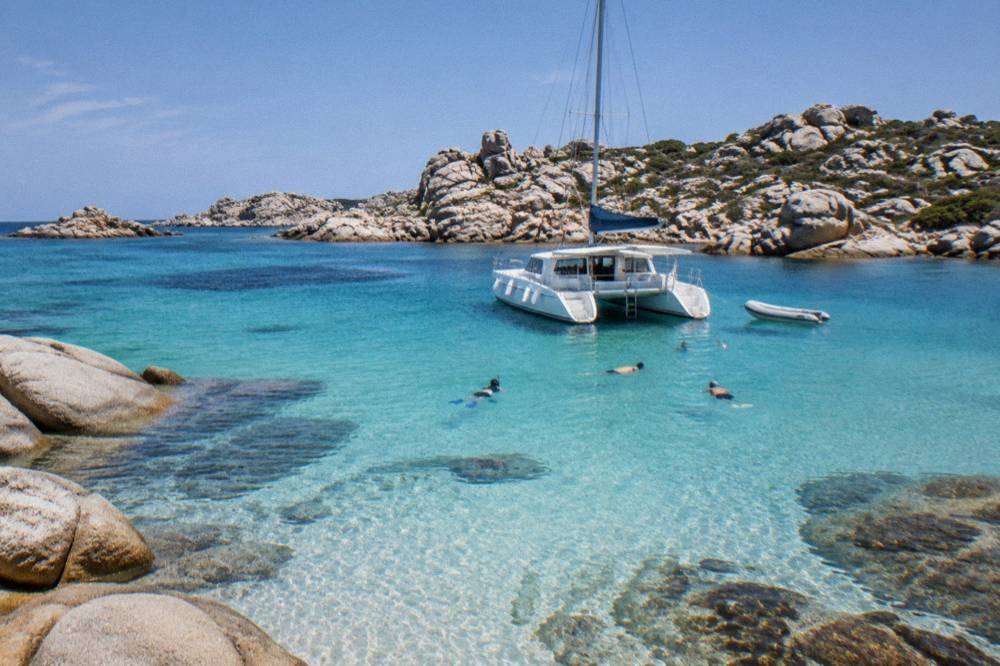 Snorkellers in the water beside a moored catamaran near a rocky Lavezzi shoreline