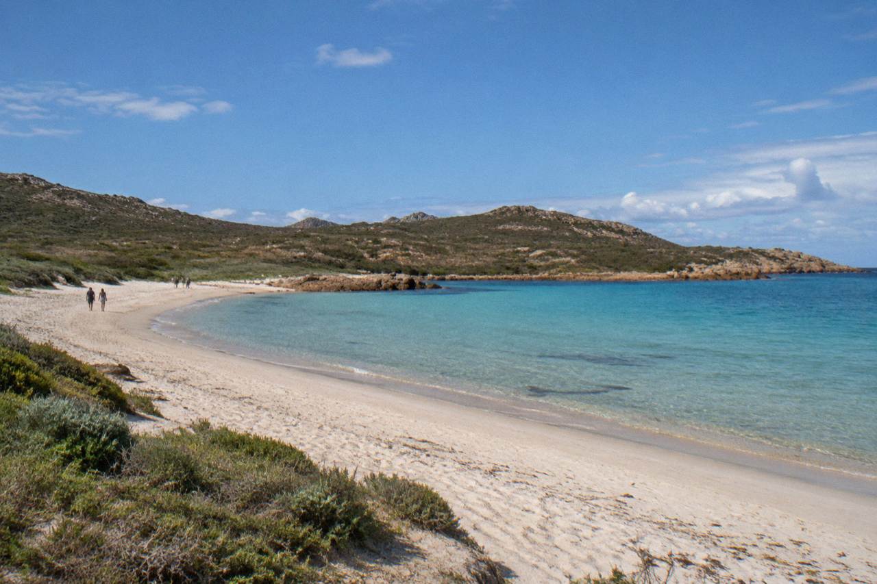 A wide empty beach of pale sand beside shallow clear water in a dry scrub landscape
