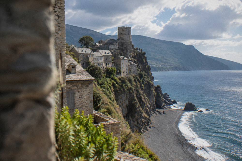 A grey village clinging to a cliff above a black pebble beach with a stone tower on top