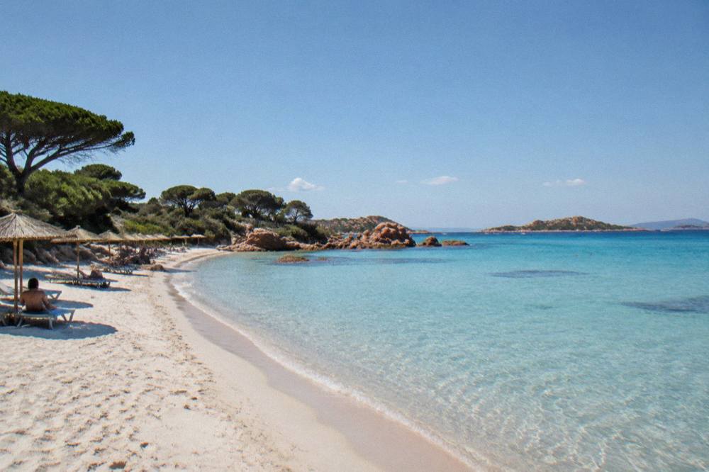 A sweep of fine pale sand backed by umbrella pines and rounded red granite boulders with shallow clear water