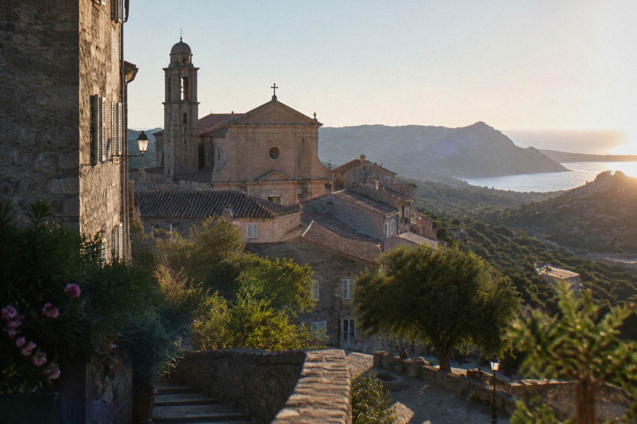 Stone buildings and a church facade above the rooftops of a Corsican hill town in warm evening light