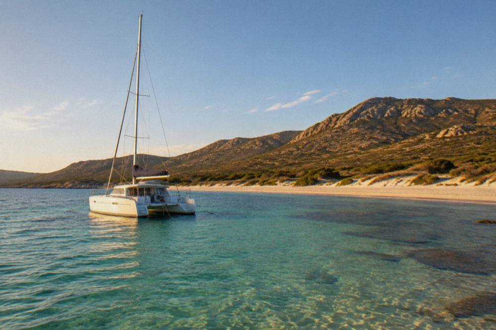 A catamaran moored in clear green water near a sandy shoreline