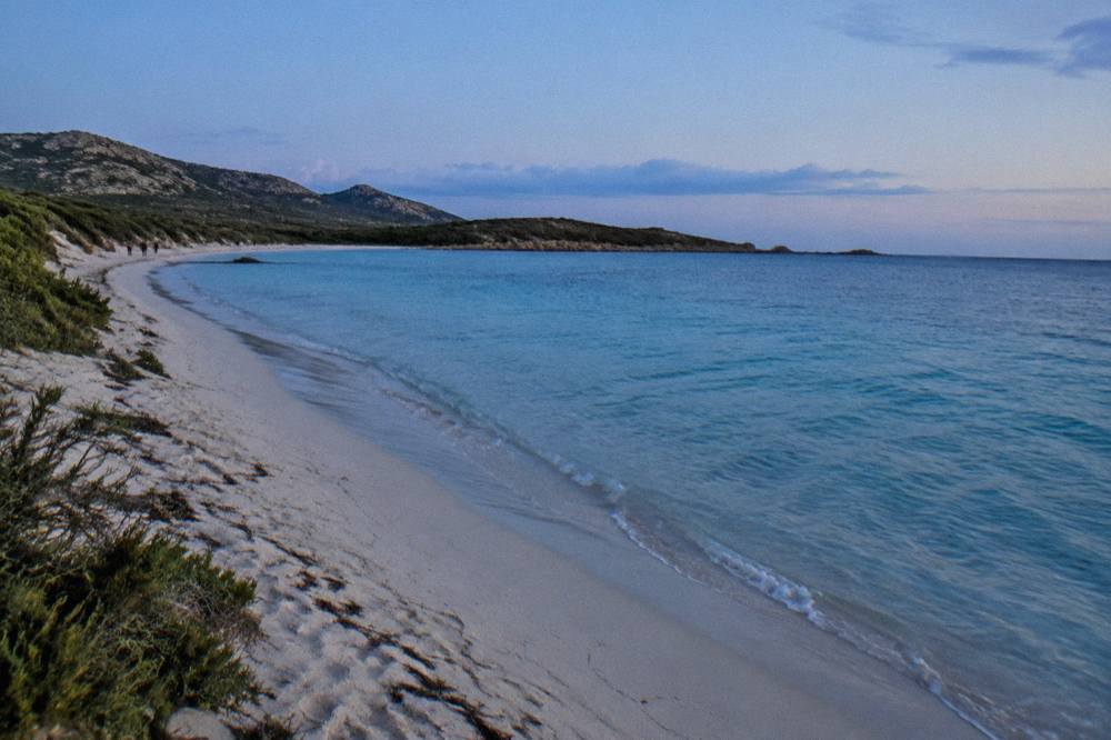 A view along a Corsican beach toward low greenheadland and open sea.