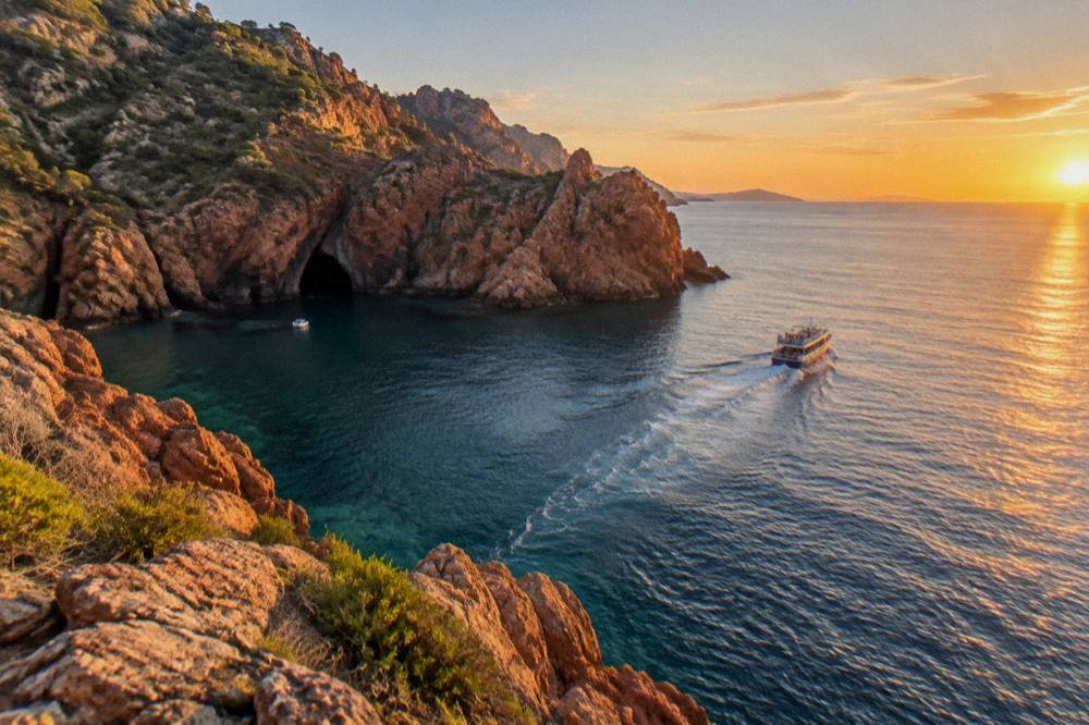 A tour boat leaving a red cliff bay toward the open sea at sunset
