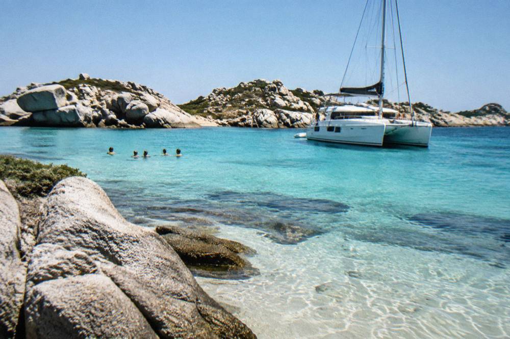A catamaran anchored near a granite island with swimmers nearby
