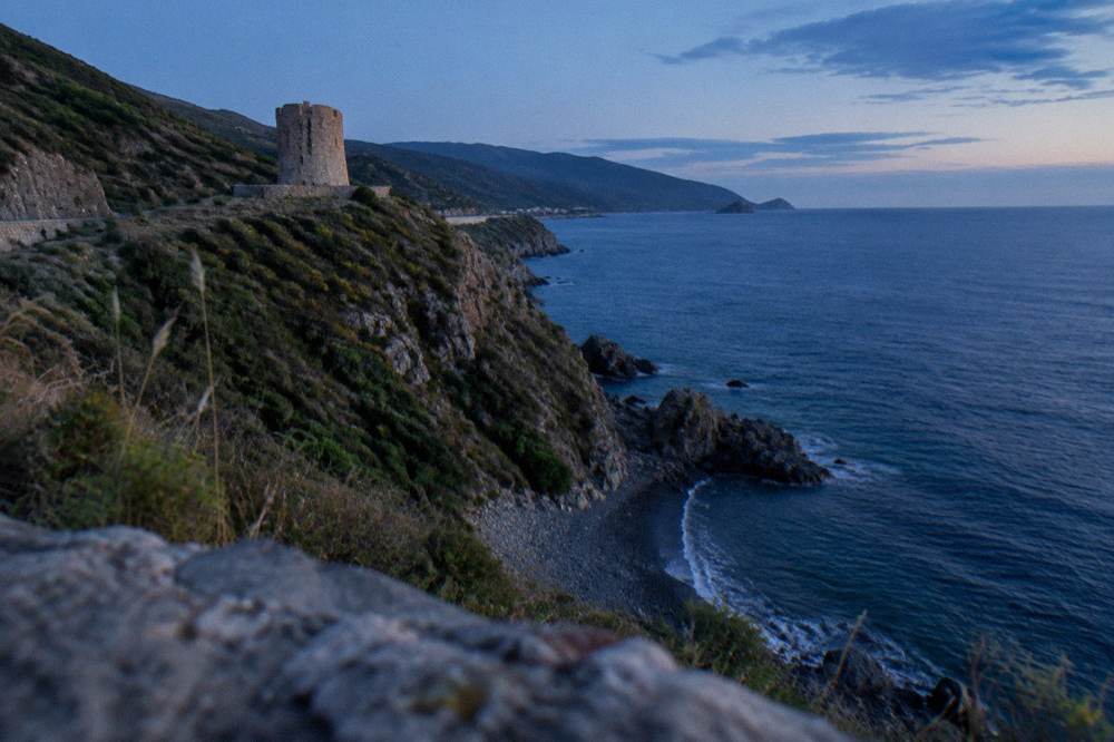 A stone tower on a grassy cliff above a dark pebble beach on the west coast of Cap Corse