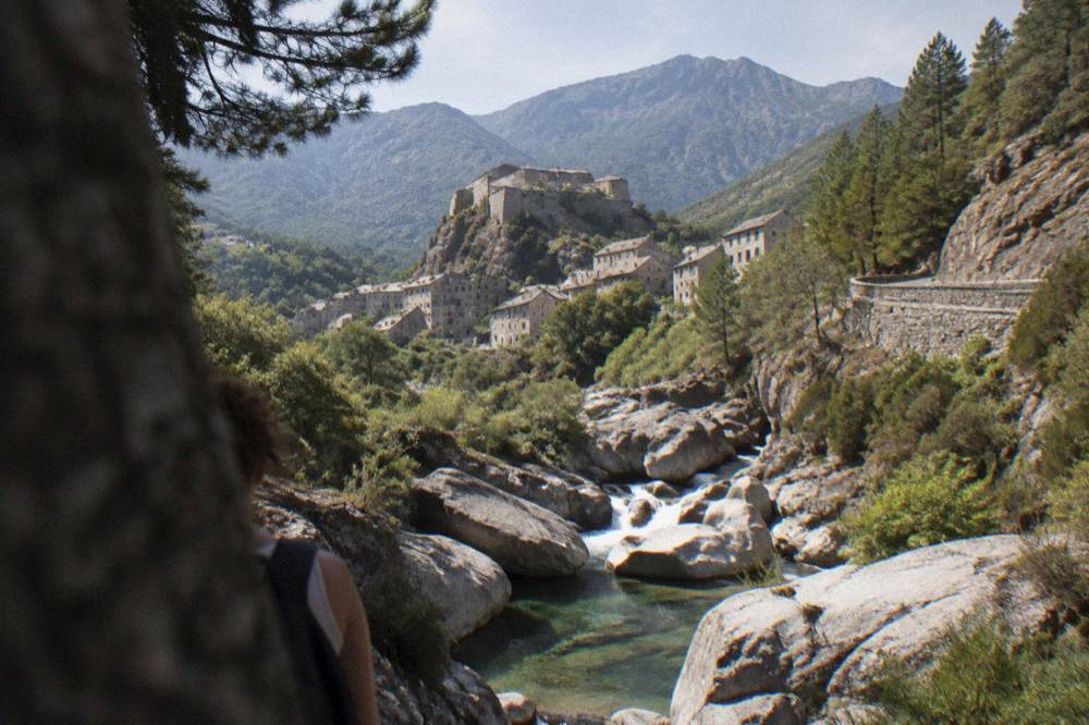 Stone town of tall grey houses below a citadel on a rock with high mountains behind