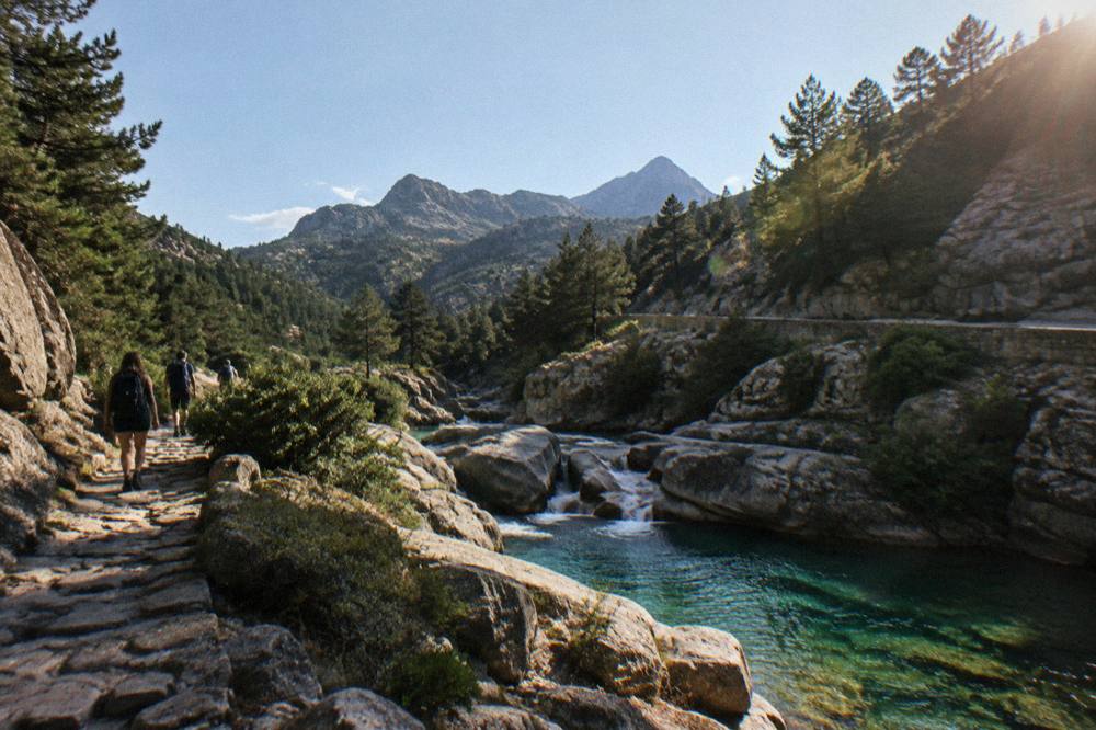 Hikers on a stone path beside a turquoise mountain lake above the Restonica valley