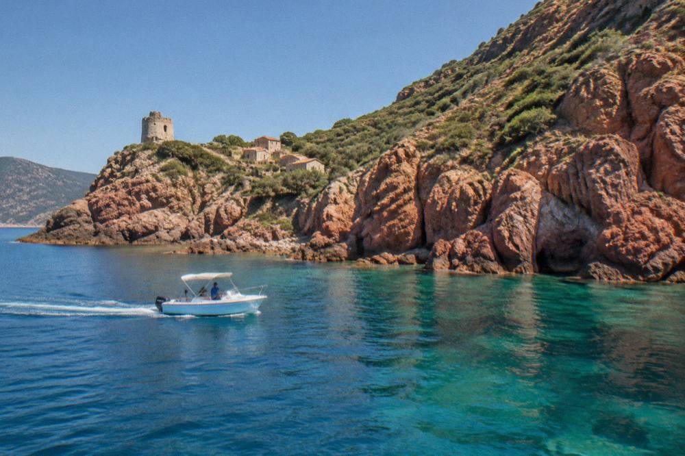 A small boat cruising close to tall red cliffs above calm sea