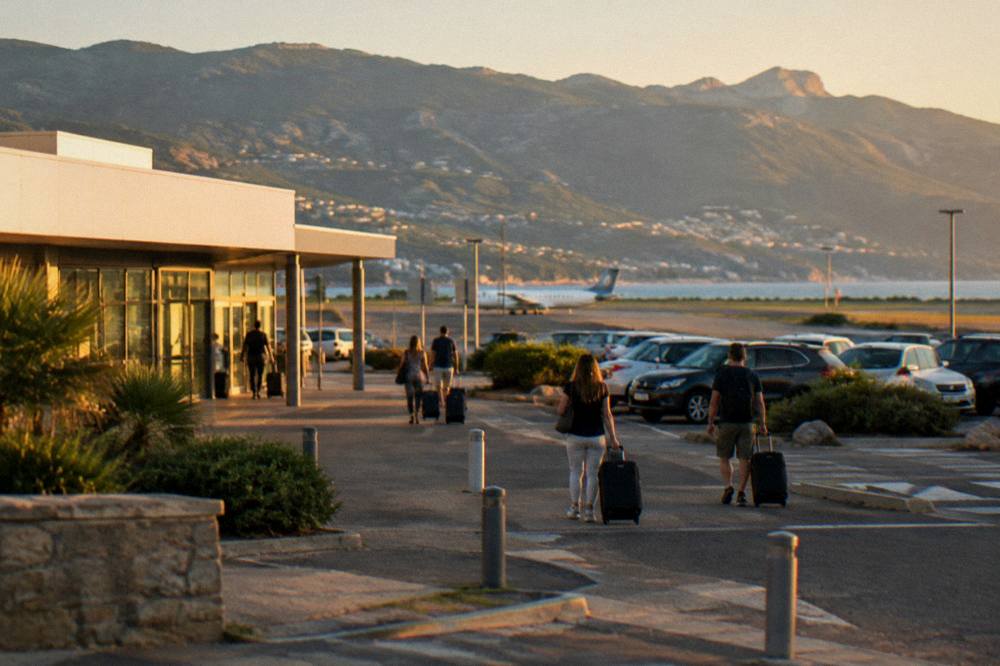 Passengers with luggage walking out of a small airport terminal toward parked cars