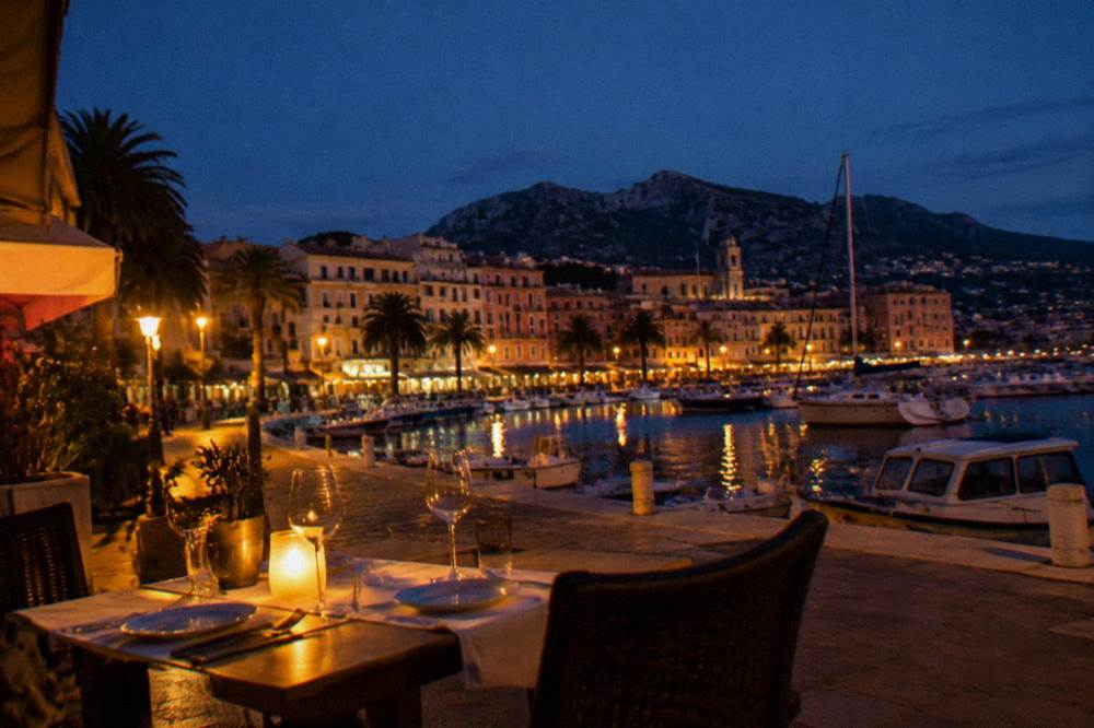A terrace table set for dinner beside a harbour at dusk with warm lights and boats behind