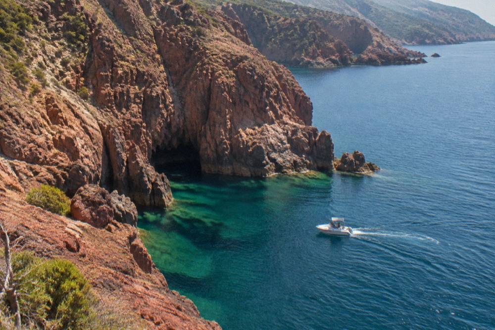 A small boat cruising close to red granite cliffs on a calm sea