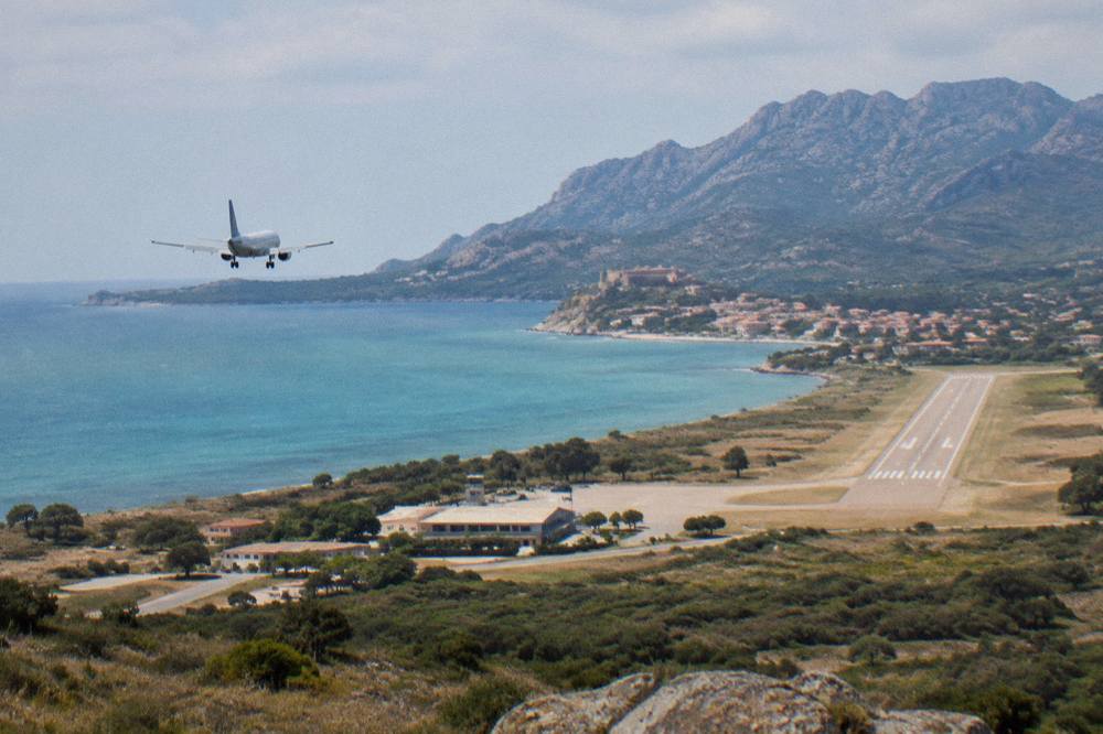 An aircraft on a low approach over turquoise water toward a runway near a Corsican coastal town