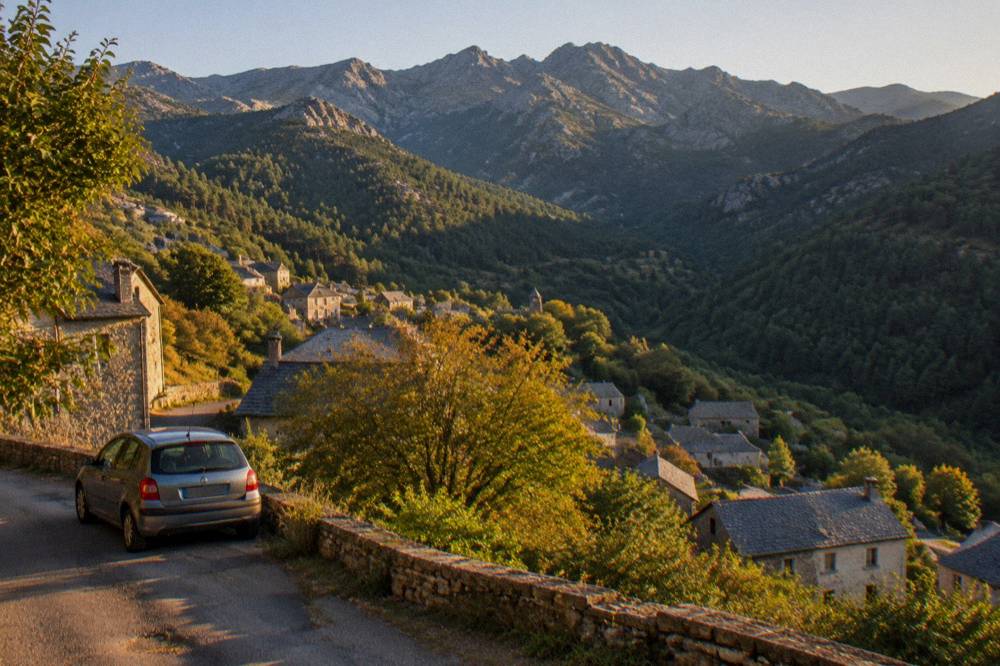 A parked small car on a narrow mountain road above hilltop villages in the Corsican countryside