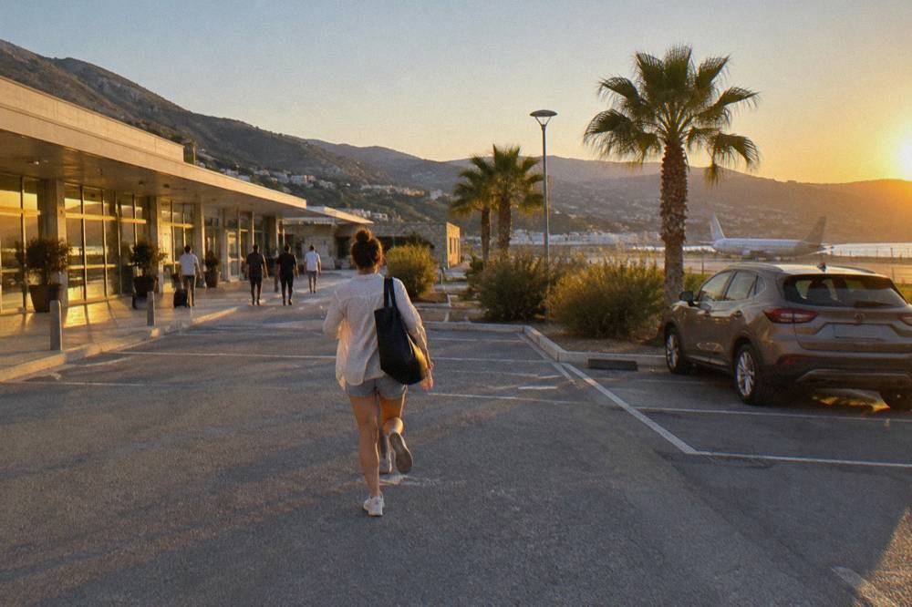 A parked rental car beside a low terminal building with hills in the distance