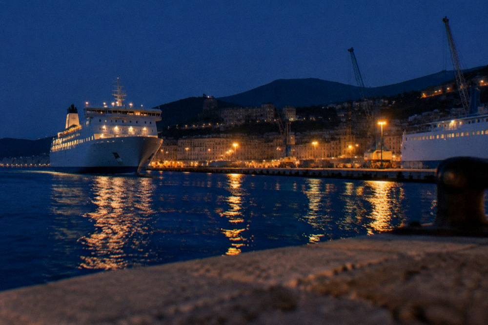 A large car ferry approaching a Corsican port town at dusk with lights along the quay