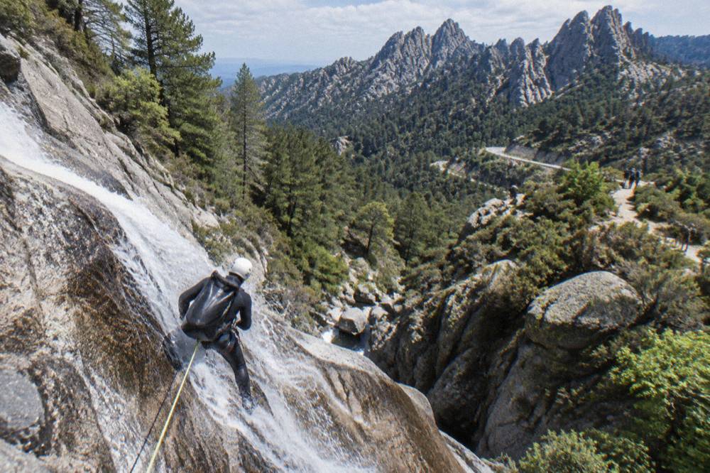 A wetsuited canyoner rappelling down a wet rock face beside a cascade in a mountain gorge