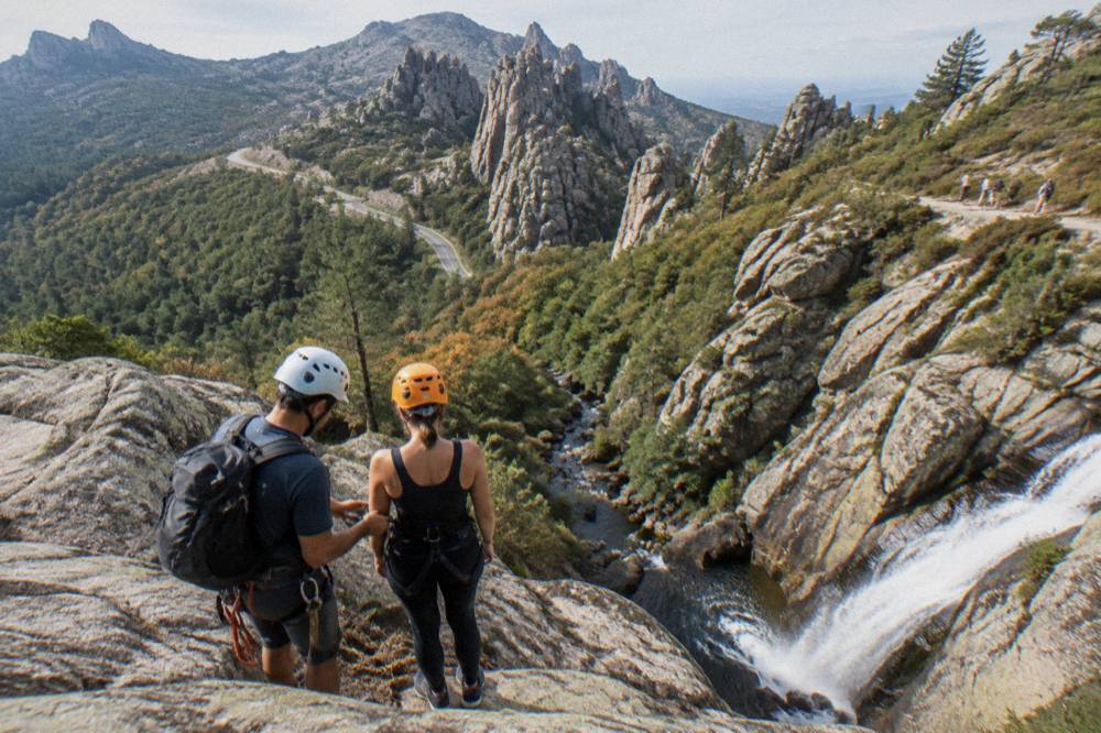 A guide checking a harness and helmet on a canyoner at the top of a rappel beside a waterfall