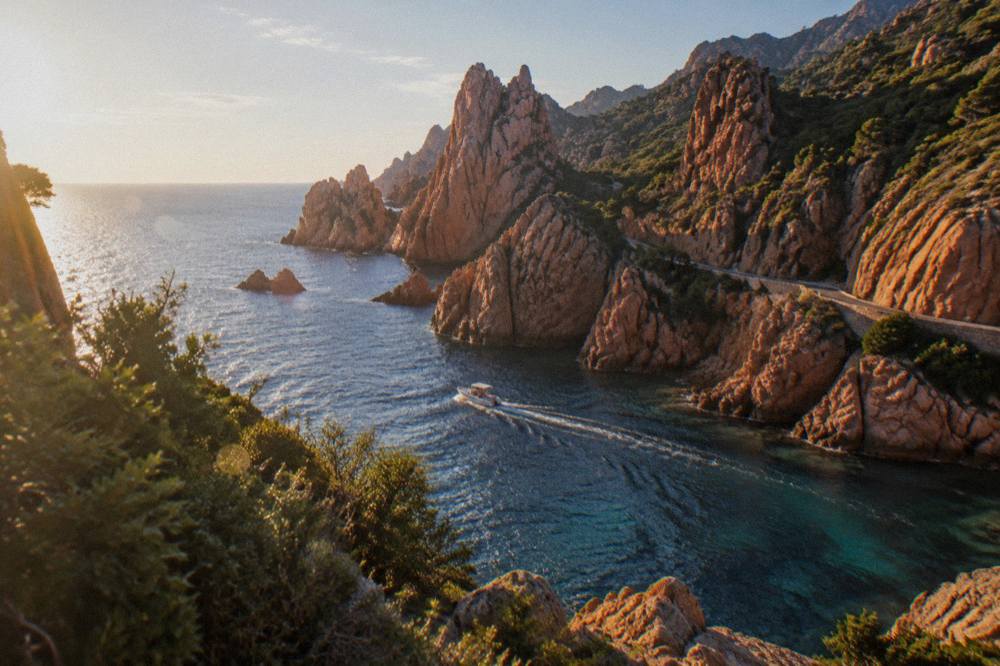 A small boat cruising along red granite cliffs above clear water on the Corsican west coast