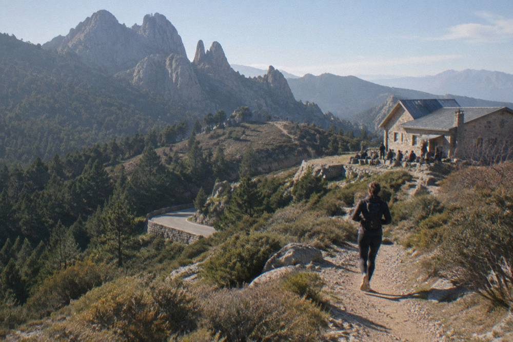A stone refuge building on a mountain ridge with hikers resting outside and peaks in the background