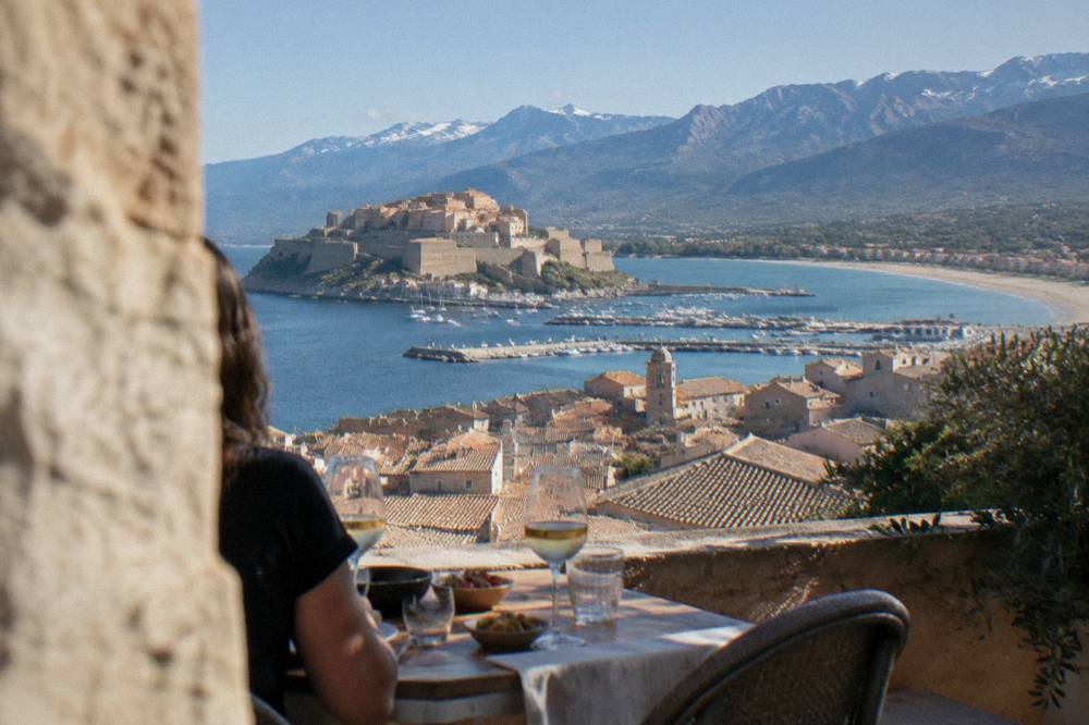 An evening table on a terrace with glasses of wine and small plates overlooking a village roofline