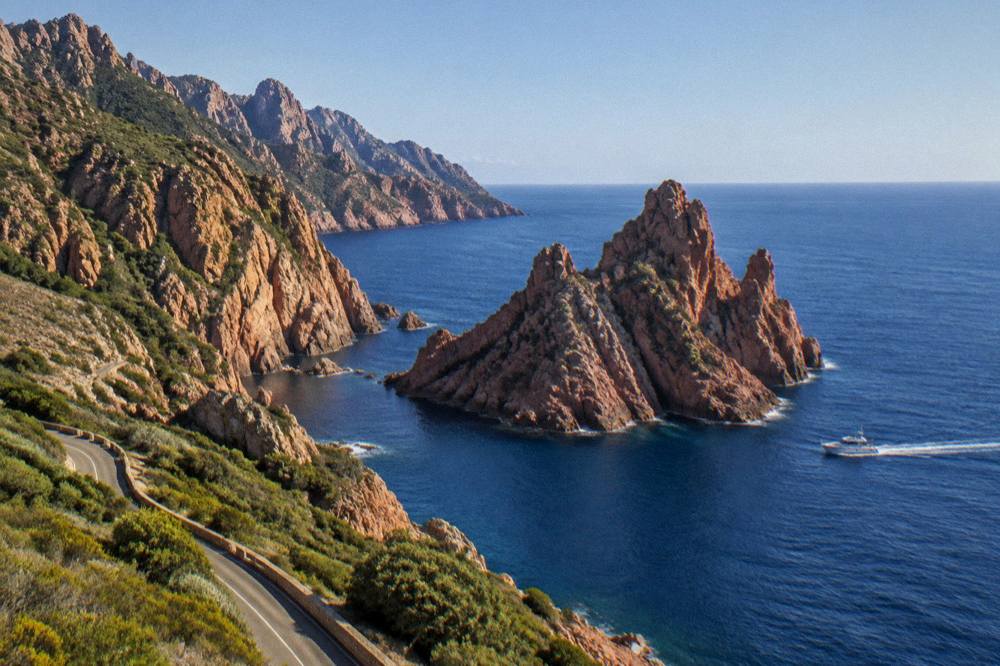 A red granite rock formation rising from the sea at the edge of a nature reserve with a boat passing below